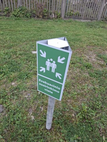 Evakuacinio susirinkimo vietos ženklas ant stovo, žalia spalva su simboliais